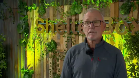 Shelterbox A man with short grey hair, glasses and a blue zipped-up top stands in front of a wooden, illuminated Shelterbox logo surrounded by plants.