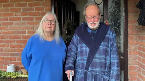 Jan and Graham stand side by side on their doorstep, looking into the camera. Jan, on the left, is wearing a bright blue v-neck sweater, and has long light grey hair and dark-framed glasses. Graham is wearing a checked blue dressing gown with a blue top, and has short dark grey hair and a white beard. He's also wearing dark-framed glasses, and is leaning on a stick.