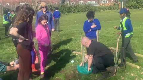 A group of children watch as a man dressed all in black takes a sapling out of a green bag full of soil.