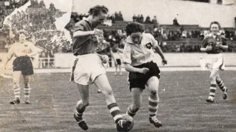 Marlene Cook Marlene Cook is pictured kicking a ball as an  opposition player attempts to make a tackle during a match. 