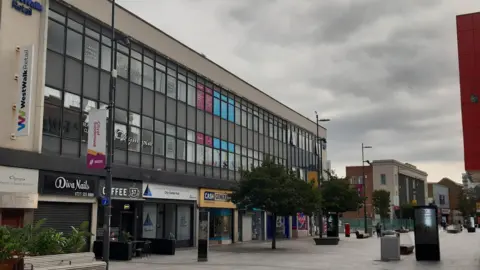 LDRS High Street West is a pedestrian street with benches and young trees in the middle. A three-storey building with many windows takes up a large section of the street. There are coffee shops, a nail and beauty salon and other units. One person is walking further down the street. 