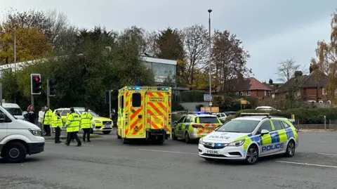 Three police cars block a busy road following a collision. An ambulance can be parked among them with its emergency lights flashing. Others cars attempting to move around the emergency service vehicles can be seen.