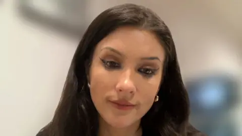 A woman with long brown hair looking at the camera. She is wearing a black top.