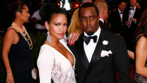 Cassie, in large diamond dangly earrings and a white gown, puts her arm on the shoulder of Sean Combs, who wears a black tuxedo with a black bowtie and white pocket square. 