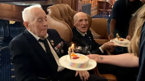 Len Chivers and Robin Boodle are seated wearing suits and ties with military medals, while staff from the charity present them with birthday cakes. 