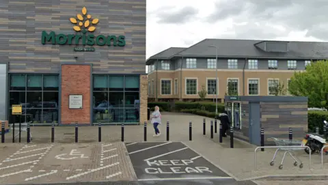 A Google Street view image of a Morrisons which the building finishes half way on the image, and is on the left. On the right is a small building with two ATM machines. A person is standing at one of the machines.