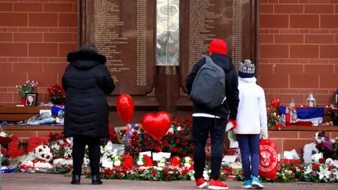 Reuters People stand in front of the Hillsborough Memorial outside Anfield Stadium in Liverpool. There are flowers and heart balloons in front of a plaque with the names of the victims of the disaster.