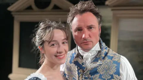 A woman in an old-fashioned dress smiling and a stern-looking man in a patterned waistcoat