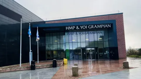 BBC Brick and glass building with sign wording saying 'HMP & YOI Grampian', and flagpoles at the side.