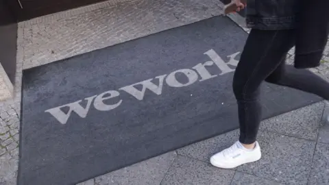 A person in black jeans and white sneakers crosses a wework door mat