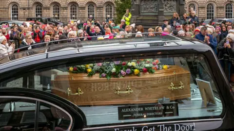PA Media A hearse with the coffin of Scottish comedian Janey Godley in it, travelling along the Royal Mile. Dozens of people can be seen watching it pass by.