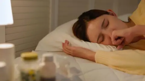 Getty Images Woman lying in bed with her eyes closed. She is coughing into her hand. There is a lamp beside the bed and what appears to be medicine.