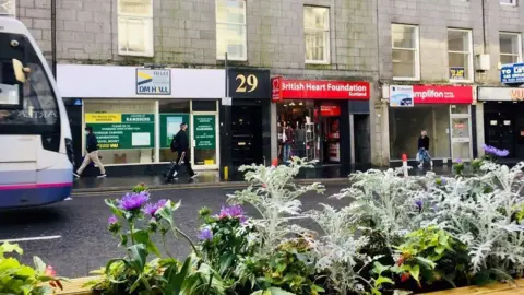 BBC Shops on Aberdeen's Union Street, some with "To let" signs, people walking on the pavement, a bus coming into shot, and flowers in the foreground.