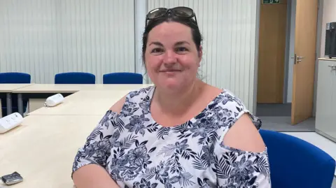 Emma Howgego/BBC Heather Williams smiles at the camera. She wears a white top with a navy flowery pattern on it and sunglasses on her head. She sits on a blue office chair next to a beige table.