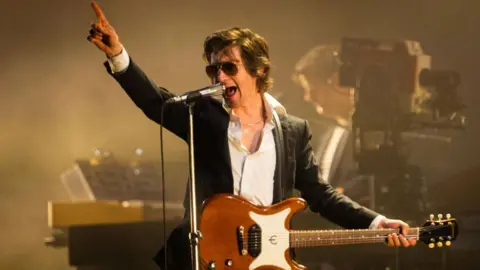 Frontman Alex Turner plays guitar on stage and sings into a microphone. He has brown hair and wears sunglasses and black blazer over a white shirt.