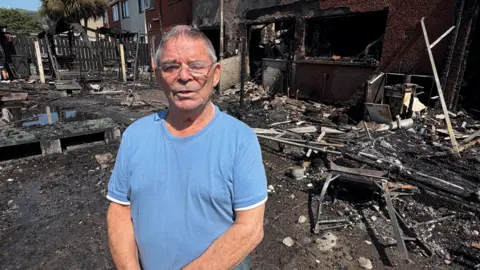 An elderly man stands outside his house