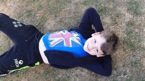 Family photo Lennix Sutcliffe pictured lying on his back on grass, with his hands behind his head, smiling at the camera. He wears a dark hoodie over a blue t-shirt with the union jack on it, and black and green jogging bottoms with the Xbox logo on them. 