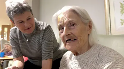Two women, one with shoulder length grey hair and a white knitted jumper and another leaning over her with a grey top and short grey hair