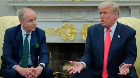 Getty Images President Donald Trump and Taoiseach Micheal Martin sitting in the Oval Office. President Trump has his arms open in a red tie and suit, while Micheal Martin has his hands clasped together