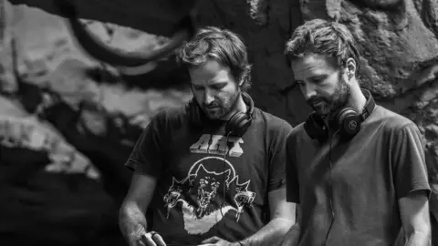 Netflix A black and white photo of the Duffer Brothers, Matt and Ross Duffer. They are both wearing headphones around their neck and are looking down. They are standing in a cave-style set.  