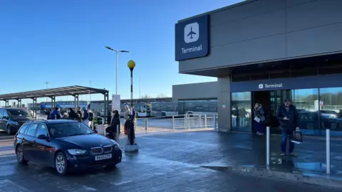 Bristol Airport drop-off parking, with a car in the foreground and people queuing up behind. Signs for the terminal can be seen at the top