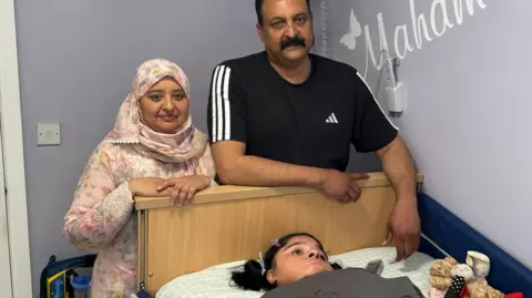 Maham lies in bed. Her parents stand close to her. Her mother wears a light floral headdress and dress while her father wears a black T-shirt with white piping.