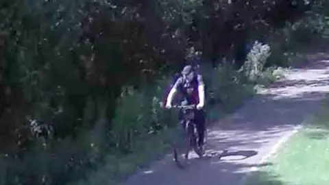 Lincolnshire Police CCTV footage showing a man on an ebike he is wearing a dark t-shirt and trousers and has a dark blue or black baseball cap on. The man is cycling on a path on one side are shrubs and trees and on the other is grass.