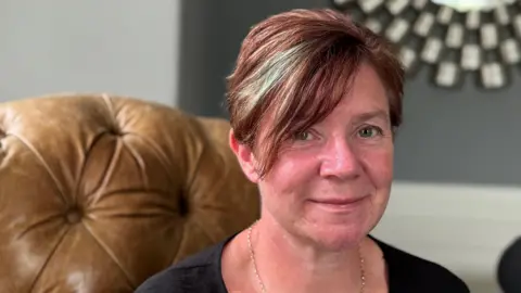 The image shows a woman sitting on a brown, tufted leather chair. They have short hair with some highlights and are wearing a black top and a thin chain necklace. In the background, there is a wall with gray paint and white trim, as well as part of a circular mirror with an intricate frame.