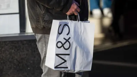 Man in dark winter jacket and grey jeans carries white paper bag with M&S logo on the side 