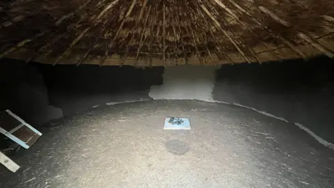 TOM JACKSON/BBC The inside of the replica roundhouse. There is straw on the floor and a square in the centre covered in ashes. The walls are made of unpainted clay and the underside of the thatched roof can also be seen. 
