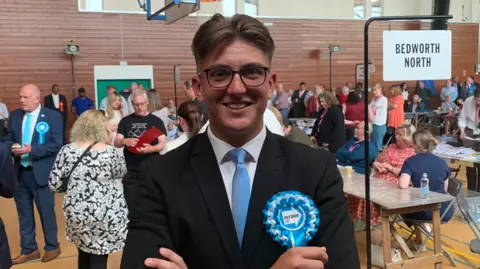 George Finch is standing in front of the count. He is wearing a jacket and tie and blue rosette and is looking happy.