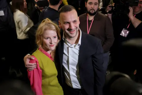 Zack Polanski celebrates Hannah Spencer's by-election victory. He is wearing a dark suit jacket with a pale open-collared shirt