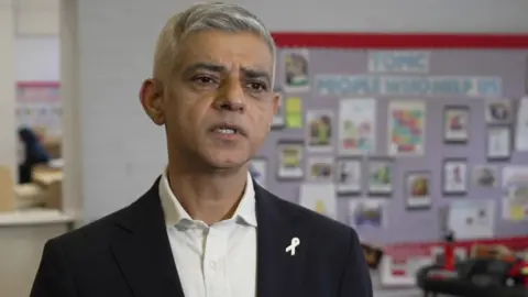 Sadiq Khan is pictured during an interview wearing a dark suit and standing in a classroom.