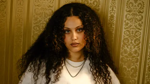 Jessy has long dark curly hair. She has a white t-shirt on and wears a gold chain.