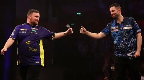 Getty Images Luke Littler and Luke Humphries stand side by side during a darts match in Sheffield. Both men are wearing short-sleeve blue tops. Littler has darts in his left hand while Humprhies gives him a thumbs up sign