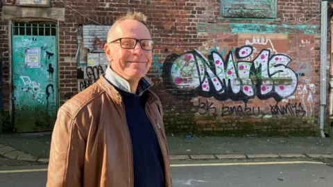 BBC/Victoria Scheer Stocks stands in front of an old brick building with worn paintwork and visible graffiti. The building’s exterior is made of dark red bricks, and it features several turquoise-painted doors and window frames that are weathered and peeling. Two upper windows have mismatched panes, and the paint on both the frames and surrounding brickwork is noticeably chipped.