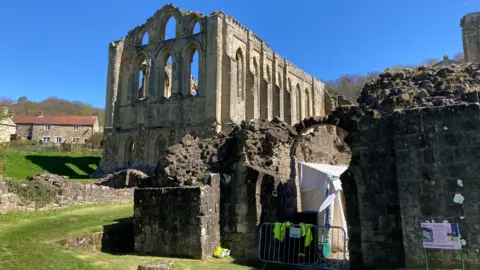 BBC / Jack Hadaway-Weller The stone ruins of a historic abbey with tall arched windows and weathered walls under a clear blue sky. In the foreground, broken stone structures and a small fenced area with equipment suggest ongoing maintenance or restoration work. Surrounding the site are grassy grounds, a few traditional houses, and bare trees, creating a peaceful rural setting.