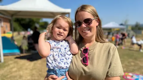 A young woman is pictured holding a little girl in what looks like a fair or festival setting. There are gazebos behind the pair. The woman has long blonde hair and is wearing a khaki T-shirt and sunglasses. She has the little girl's sunglasses - which are pink with white dots - hanging from the neckline of her T-shirt. The little girl is wearing a blue and white summer top and denim shorts.