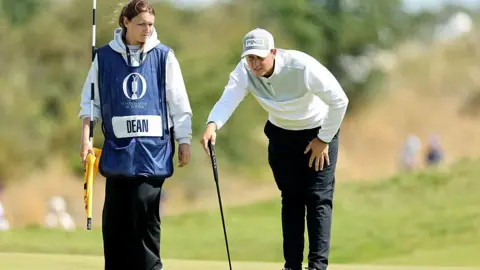 Getty Images A golfer in a grey jumper and cap bends down to study the green while his female caddie holds the flag.