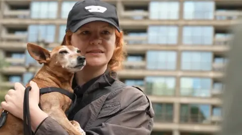 A woman with short ginger hair wearing a cap, holding a small dog in front of a block of flats