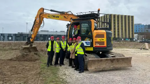 Nikki Fox/BBC Andrew Stephenson and NHS staff in front of a digger