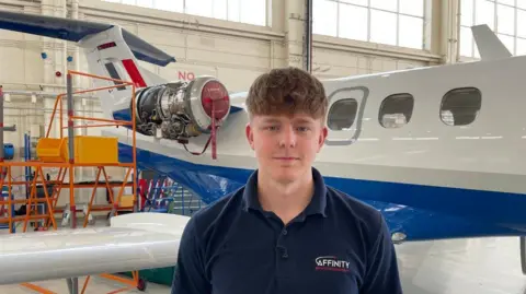 A young man with brown hair draped over his forehead who is clean shaven is smiling at the camera and wearing a navy polo shirt which says Affinity on it. Behind him is a small aircraft which is white, blue and red, which is stationary inside a large warehouse building.