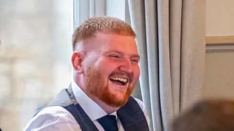 Megan Ahmed Picture shows a man with ginger hair and a moustache and beard wearing a blue short, tie and waistcoat, sat down laughing