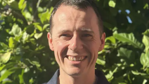 Dr Matthew Cook A man with short brown hair is smiling at the camera. There is a hedge behind him.