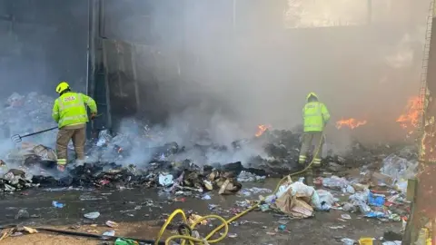 Two firefighters are at a waste recycling centre. One of them is holding a hose and the other is holding a pitch fork. They are both wearing firefighter uniform including a yellow fluorescent helmet and yellow fluorescent jackets which says FIRE & RESCUE on the back.