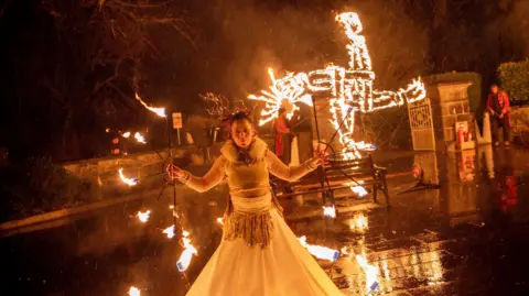 Kildare County Council/Allen Kiely A woman dressed in festival costume stands in front of a large, burning St Brigid's cross, illuminating the night sky as part of festival celebrations in Maynooth