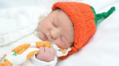 Newborn baby wearing white cardigan, orange pumpkin hat and clutching white blanket with orange pumpkins on it. He is laid down asleep on a white sheet and has his eyes scrunched shut.