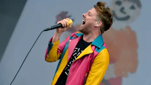 A picture of the singer of the Kaiser Chiefs on stage. He is wearing a pink, yellow and blue leather biker jacket and singing into a yellow and black microphone.