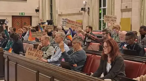 Bristol Green Party Inside a council meeting with about two dozen councillors on rows of red benches with many waving banners saying things like 'trans men are men' and Pride flags.