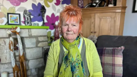 Becki Bowden / BBC Jenny Thurlow has orange hair and wears a green top and scarf as she sits in her home in front of a stone fireplace